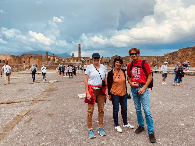Two tourists accompanied by a local guide at the Roman Forum in Pompeii, with ancient ruins and columns visible in the background under a cloudy sky